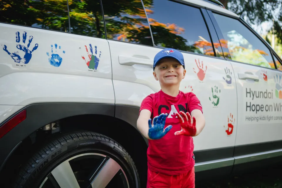Young boy in red tee shirt with a blue baseball cap holding up his hands covered in paint. Behind him is a white Hyundai with painted hand-prints all over.