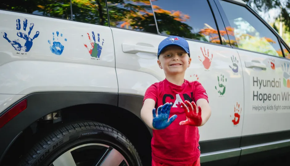 Young boy in red tee shirt with a blue baseball cap holding up his hands covered in paint. Behind him is a white Hyundai with painted hand-prints all over.